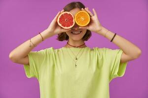 Tonårs flicka, glad och Lycklig, med brunett kort hår innehav grapefrukt och orange över henne ögon. stående över lila bakgrund. bär grön t-shirt, tänder tandställning, armband och halsband foto