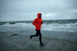 ung man med frodig skägg Träning på strand i kall tidigt morgon, bär svart sportig kläder och värma orange täcka med huva medan joggning. kondition och sport begrepp foto