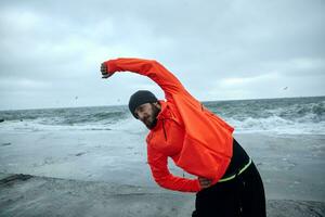bild av ung mörk håriga sportig man med skägg håller på med fysisk övningar över havet bakgrund på grå kall morgon, stretching muskler innan träna. kondition manlig modell foto