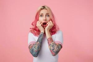 chockade Söt ung kvinna med tatueringar och rosa lockigt hår stående över rosa bakgrund med bred mun öppnad, lutande haka på Uppfostrad händer och rynkar ansikte förvånad foto