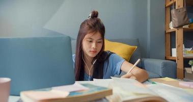 ung asiatisk tjej tonåring med slitage hörlurar använda bärbar dator lära sig online skriva föreläsning anteckningsbok i vardagsrummet hemma. isolera utbildning online e-learning coronavirus-pandemikoncept. foto