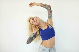 positiv ung attraktiv blond kvinna klädd i tillfällig kläder höjning henne tatuerade händer och leende angenämt med stängd ögon, isolerat över vit bakgrund foto