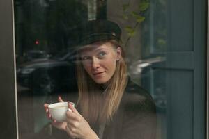 attraktiv ung blond kvinna i trendig kläder Sammanträde över Kafé interiör och ser eftertänksamt på fönster, har kopp av te medan väntar för henne beställa foto