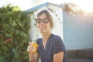 utomhus- skott av charmig ung brunett kvinna i årgång solglasögon dricka orange juice med sugrör, Sammanträde över grön bakgård och ser på kamera positivt foto