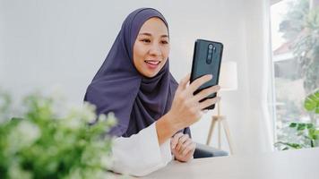 ung asiatisk muslimsk affärskvinna som använder smart telefon prata med en vän genom videochatt -brainstorm online -möte medan hon arbetar på distans hemifrån i vardagsrummet. social distansering, karantän för corona. foto