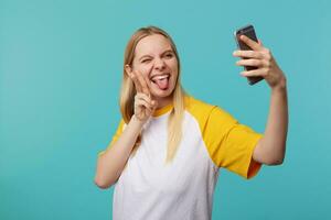 inomhus- Foto av ung Söt blond lång håriga lady förvaring henne hand Uppfostrad medan framställning selfie på henne mobil telefon, framställning roligt medan Framställ över blå bakgrund