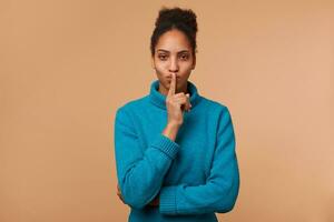 mystisk afrikansk amerikan ung lady, demonstrerar en gest av tystnad, innehav ett index finger nära de mun samtal för Integritet till vara bevarade, hemlighet, stanna kvar tyst, lugn. isolerat över beige bakgrund. foto