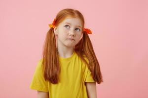 porträtt liten tänkande fräknar rödhårig flicka med två svansar, utseende bort, finputsning kinder, bär i gul t-shirt, står över rosa bakgrund med kopia Plats. foto