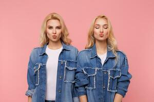 positiv attraktiv ung blond systrar med lockar hopfällbar mun i luft kyss medan stående mot rosa bakgrund och förvaring händer längs deras kroppar foto