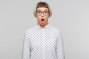 porträtt av förvånad förvånad blond ung kvinna med öppnad mun bär polka punkt skjorta och glasögon utseende chockade och skrikande isolerat över vit bakgrund foto