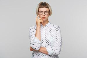 närbild av fundersam koncentrerad blond ung kvinna bär polka punkt skjorta och glasögon håller händer vikta och tänkande isolerat över vit bakgrund foto