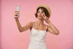 porträtt av charmig ung kvinna med kort brun hår bär vit sommar klänning och sugrör hatt, höjning hand med fred gest medan framställning selfie med henne smartphone, isolerat över rosa bakgrund foto