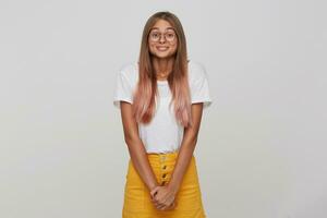 porträtt av leende förvirrad ung kvinna studerande med lång färgade pastell rosa hår bär t skjorta gul kjol och glasögon känner Lycklig och generad isolerat över vit bakgrund foto