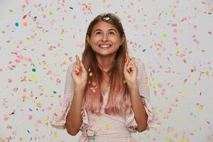 porträtt av glad upphetsad ung kvinna med lång färgade pastell rosa hår och fingrar korsade bär polka punkt rosa klänning framställning en önskar och har fest isolerat över vit bakgrund med konfetti foto