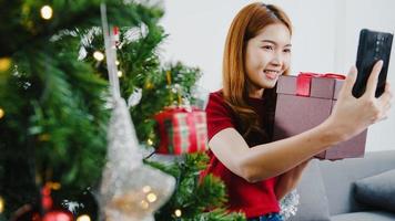 ung asiatisk kvinna som använder smarta telefonsamtal och pratar med par med julklappslåda, julgran dekorerad med prydnad i vardagsrummet hemma. jul- och nyårsfest.