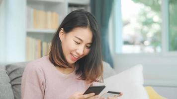 ung leende asiatisk kvinna som använder smartphone och köper onlineshopping med kreditkort medan hon ligger på soffan när hon kopplar av i vardagsrummet hemma. livsstil latin och spansktalande etnicitet kvinnor hemma koncept.