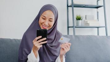 asien muslimsk dam använder smart telefon, kreditkort köper och köper e-handel internet i vardagsrummet hemma. stanna hemma, shoppa online, självisolering, social distans, karantän för coronavirus. foto