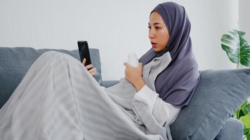 ung asien muslimsk dam bär hijab med telefonsamtal och pratar med läkarkonsultation eller onlinekonsultation på soffan i vardagsrummet hemma. social distansering, karantän för coronavirus -koncept.