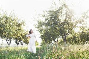 en ung flicka i en vit lång klänning går i trädgården. vacker solnedgång genom trädens löv foto