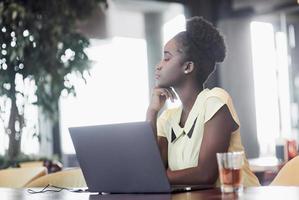 en ung afroamerikansk tjej med mörkt lockigt hår som funderar över en bärbar dator på ett café