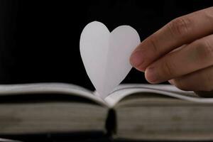 bok med öppen sidor och hjärta formad papper. kvinna hand innehav hjärta formad papper över öppen bok på tabell. kärlek av läsning begrepp foto