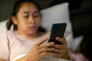 ung asiatisk kvinna liggande på vit säng och spelar smartphone på natt. kvinna i säng med telefon. hälsa och social begrepp. foto