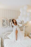 ung lockigt flicka med lång hår i lyxig rum stående på säng i vit öppen klänning innehav många gel uppblåsbar bollar i händer och framställning för bröllop ceremoni foto