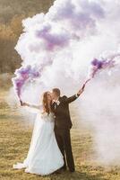 brud och brudgum spelar med färgad rök i lila händer, kramar och kyssar. rök bomber på en bröllop. foto