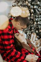 ung skön par i matchande röd pyjamas nära de jul träd. glädje, kramar och kyssar. ny år högtider och gåvor under de jul träd foto