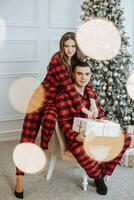 ung skön familj par i identisk röd pyjamas nära de jul träd glädjas och utbyta jul gåvor i deras händer. ny år högtider och gåvor under de jul träd foto