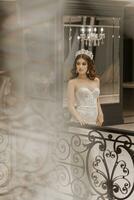 studio Foto av en flicka i en bröllop klänning nära en svart metall räcke. en stor hängande kristallkrona. lyx interiör i vit stil