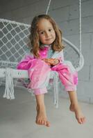 porträtt av en Lycklig liten flicka i rosa pyjamas på de veranda, liggande på en korg- gunga och spelar med henne liten leksak foto