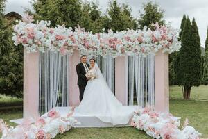 attraktiv brud och brudgum på de ceremoni på deras bröllop dag med ett båge tillverkad av rosa och vit blommor. skön nygifta, en ung kvinna i en vit klänning med en lång tåg, män i en svart kostym. foto