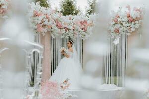 attraktiv brud på de ceremoni på henne bröllop dag med ett båge tillverkad av rosa och vit blommor. skön nygifta, en ung kvinna i en vit klänning med en lång tåg, män i en svart kostym. foto