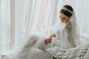 en skön flicka med en bröllop frisyr och en tiara på henne huvud i en transparent morgonrock är framställning för en bröllop i en hotell med en kunglig interiör. lång slöja, naturlig ljus, porträtt Foto