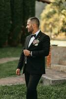 stilig skäggig man brudgum i svart kostym och svart rosett slips stående utomhus. bröllop porträtt. en man i en klassisk kostym foto