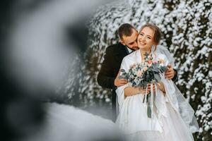 känslig porträtt av Lycklig nygifta. de brudgum kramar de brud i de vinter- parkera. leende brud i bröllop klänning och vit poncho. de brudgum är klädd i en svart täcka. bred vinkel. fri Plats foto