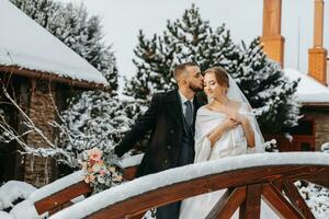 Lycklig brud och brudgum omfamning bland snöig träd på en trä- bro. brudgum och brud i de vinter- parkera. brud med en bukett av blommor i en bröllop klänning och poncho. brudgum i en svart täcka. foto