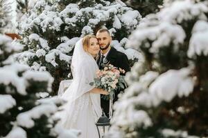 porträtt av de brud och brudgum mellan snöig träd. de brudgum kramar de brud i de vinter- parkera. brud med en bukett av blommor i en bröllop klänning och poncho. de brudgum är klädd i en svart täcka. foto