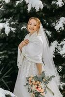 en leende och Lycklig brud på de bakgrund av snöig jul träd med en frodig bukett av blommor i henne händer. porträtt av en skön brud i en vit poncho och bröllop klänning i en vinter- parkera foto