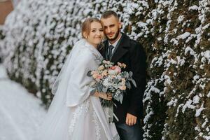 porträtt av Lycklig nygifta på de bakgrund av snötäckt träd. de brudgum kramar de brud i de vinter- parkera. leende brud i bröllop klänning och vit poncho. de brudgum är klädd i en svart täcka. foto
