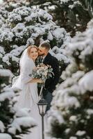 porträtt av de brud och brudgum mellan snöig träd. de brudgum kramar de brud i de vinter- parkera. brud med en bukett av blommor i en bröllop klänning och poncho. de brudgum är klädd i en svart täcka. foto