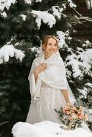 en leende och Lycklig brud på de bakgrund av snöig jul träd med en frodig bukett av blommor i henne händer. porträtt av en skön brud i en vit poncho och bröllop klänning i en vinter- parkera foto
