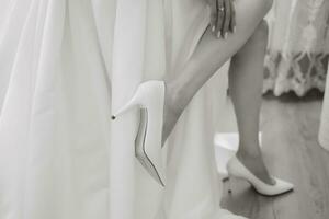 skön modell flicka med lång lockigt hår i en modern bröllop klänning. vertikal porträtt. en skön brud i en vit bröllop klänning bär högklackade skor. svart och vit Foto
