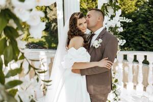 ett elegant brud och brudgum utgör tillsammans utomhus på en solig bröllop dag mot en bakgrund av blommor i en skön plats. de brudgum försiktigt kramar de brud och kyssar henne. foto