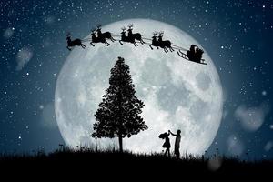silhuetten av jultomten får ett drag att rida på sina renar över fullmåne vid nattjul. njuter av par som dansar under fullmåne. foto