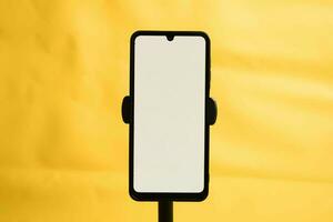 porträtt telefon med vit skärm fast till stativ på gul bakgrund, för attrapp design. foto
