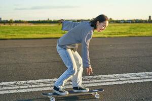 Lycklig skater flicka ridning henne skateboard och har roligt på tömma gata. leende kvinna njuter kryssare rida på solig väg foto