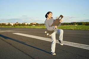 Lycklig brunett asiatisk flicka har roligt på gata med longboard, skridskoåkning, använder sig av skateboard som gitarr, njuter skateboard foto