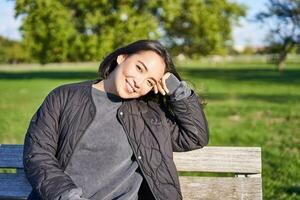 skönhet porträtt av leende asiatisk flicka med mörk kort hår, Sammanträde på bänk i parkera och gazing på kamera foto